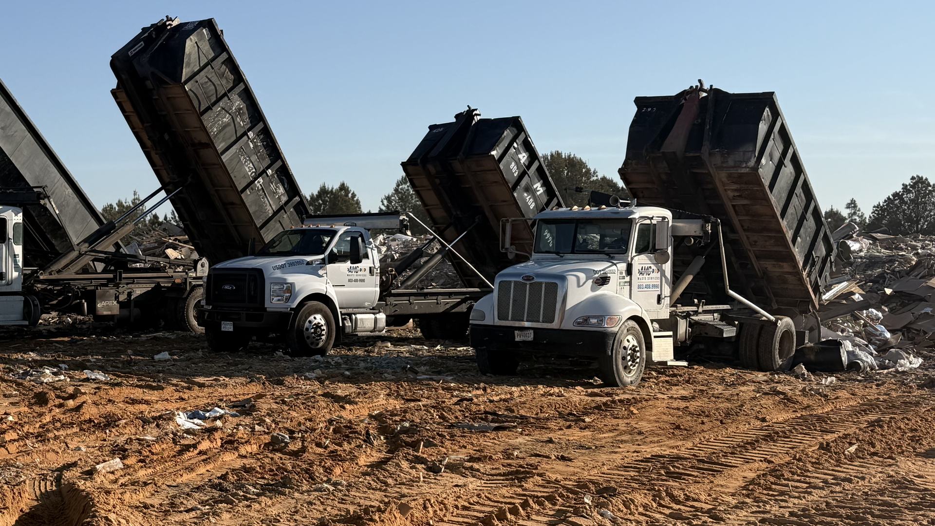 Three AMC Waste Services trucks unloading roll-off dumpsters with their beds raised