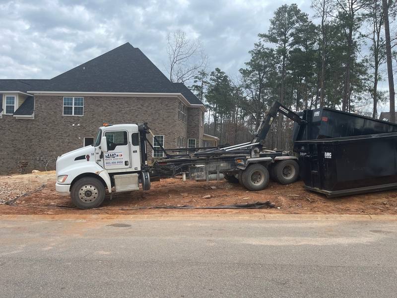 An AMC truck setting a roll-off dumpster in the driveway of a brick house
