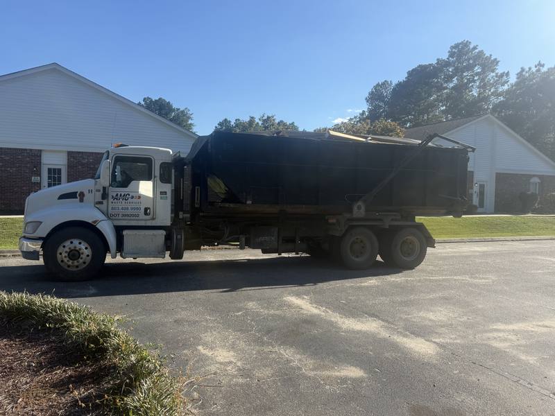 An AMC Waste Services truck picking up a roll-off dumpster at a commercial job
